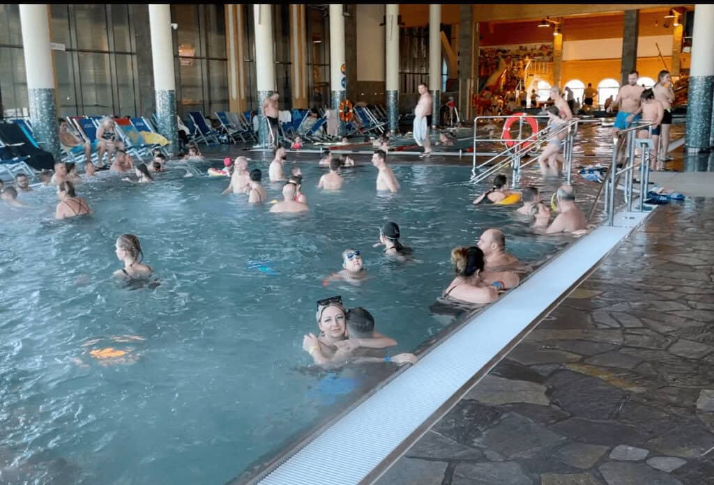 Indoor pool with many people swimming and relaxing in the water at Zakopane's renowned thermal baths; lounge chairs line the side and pool stairs provide access.