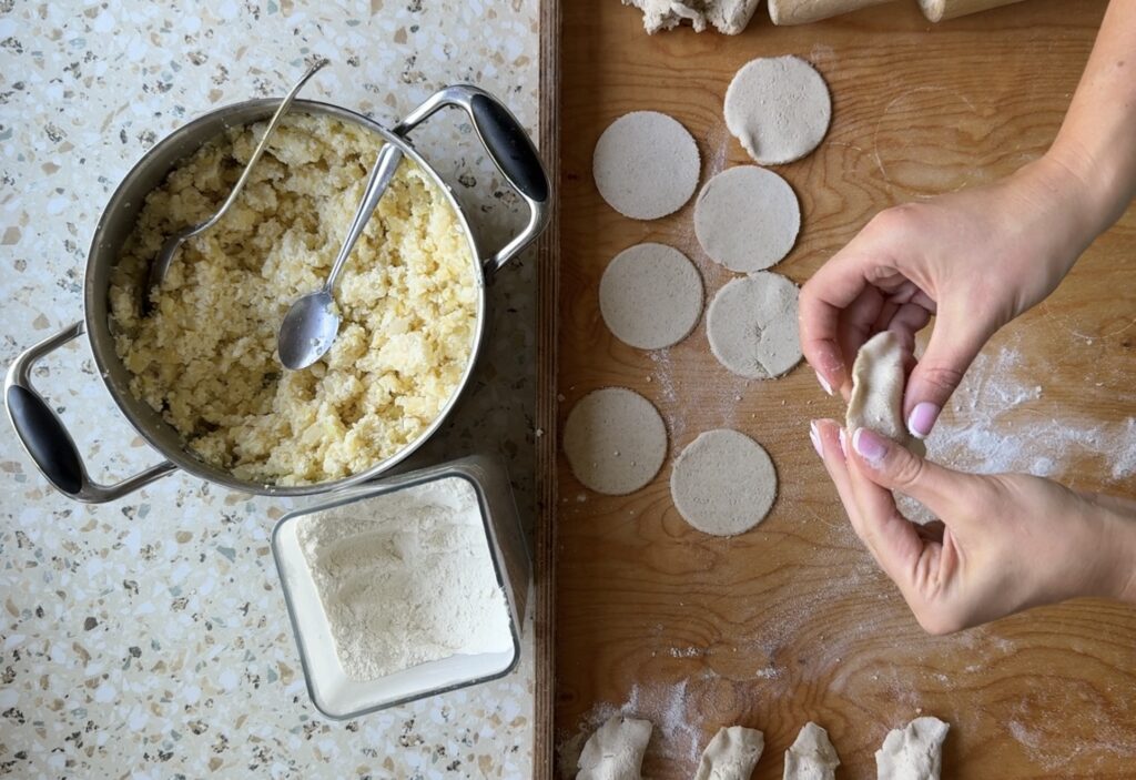 Hands folding dough circles with filling next to a pot of mixture and a container of flour on the countertop&mdash;just like making Authentic Pierogi in Babcia's kitchen.
