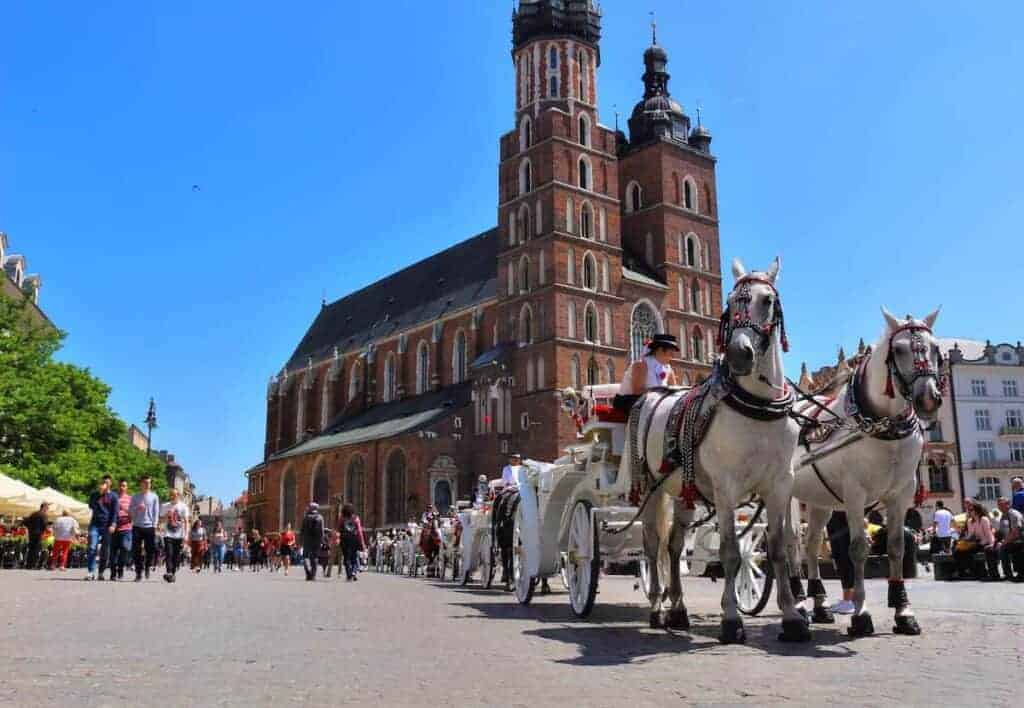 A white horse-drawn carriage passes in front of a large brick church with twin towers on a sunny day, as people stroll nearby&mdash;capturing the charm of a Poland city often counted among the top cities Poland has to offer.