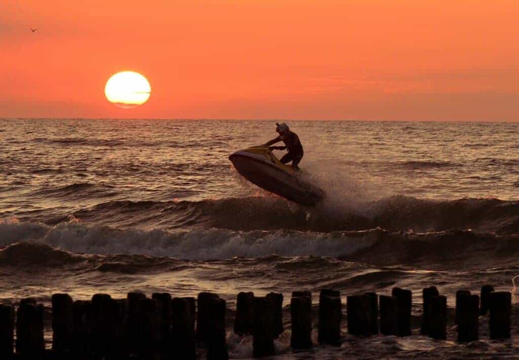 A person rides a jet ski over waves near wooden posts during sunset in Zachodniopomorskie, one of the best places to visit in Poland, with the sun low over the horizon.