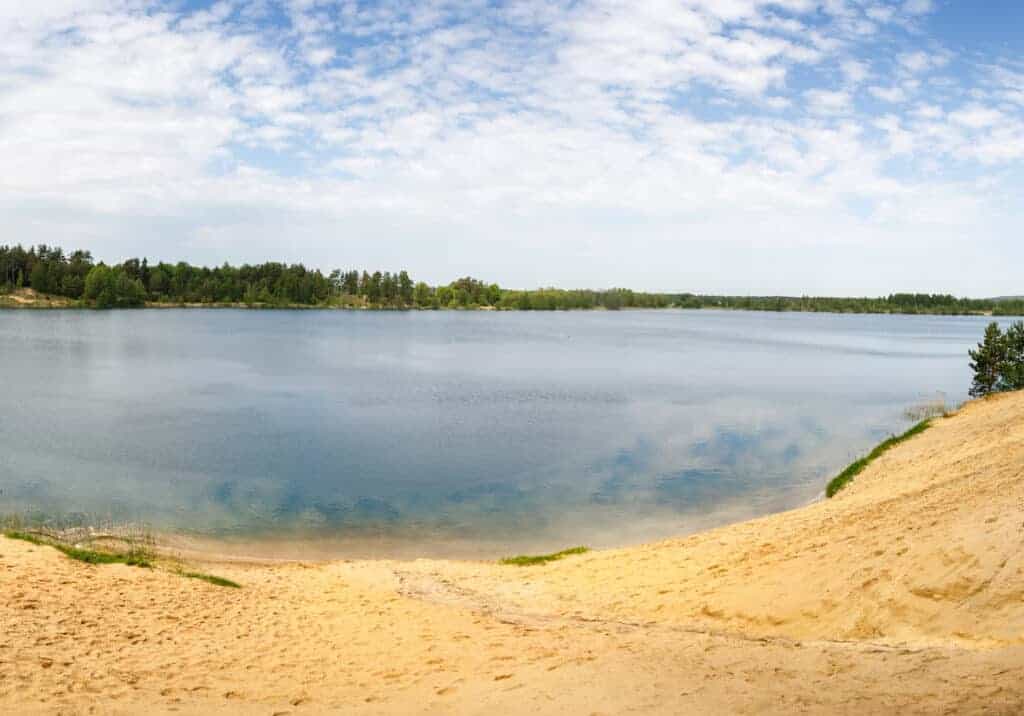 A calm lake bordered by sandy shores and forested trees under a partly cloudy sky, reminiscent of the peaceful beaches in Krakow.
