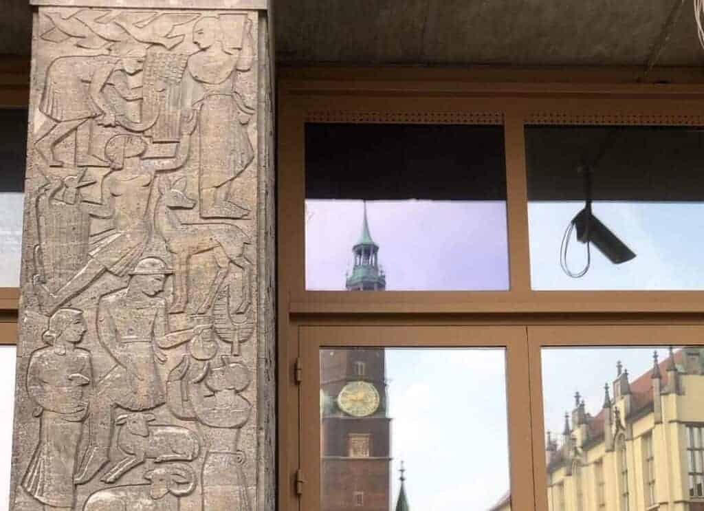 Stone pillar with carved figures on the left, and glass windows reflecting a clock tower and nearby buildings on the right. A camera is mounted above the window, capturing the essence of Street Art Wroclaw in this vibrant urban scene.