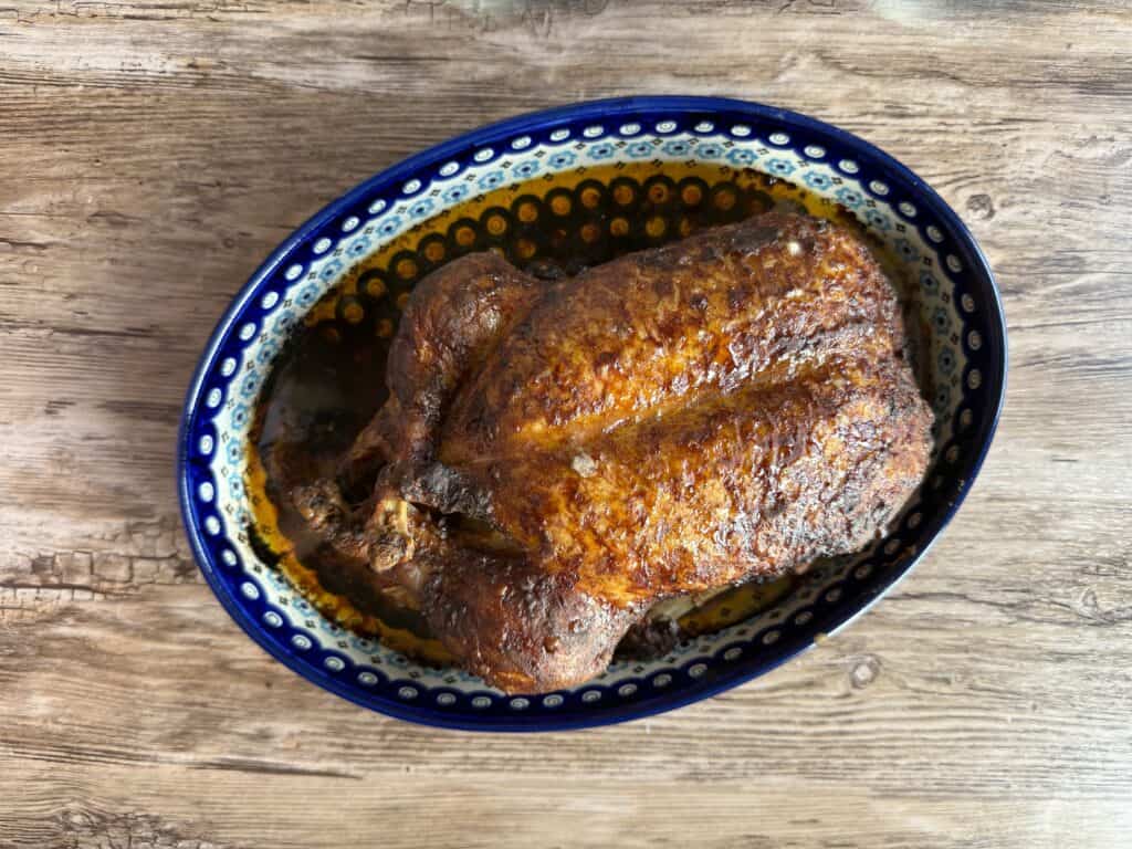 A whole roasted chicken with a crispy, seasoned skin sits in a decorative oval ceramic dish on a wooden surface, reminiscent of a traditional Polish duck recipe presentation.