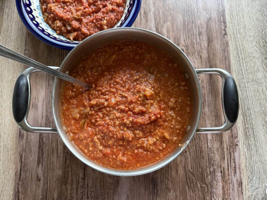A pot of chunky tomato-based Polish stuffed cabbage soup with a ladle sits next to a bowl containing a similar soup, all on a wooden surface.