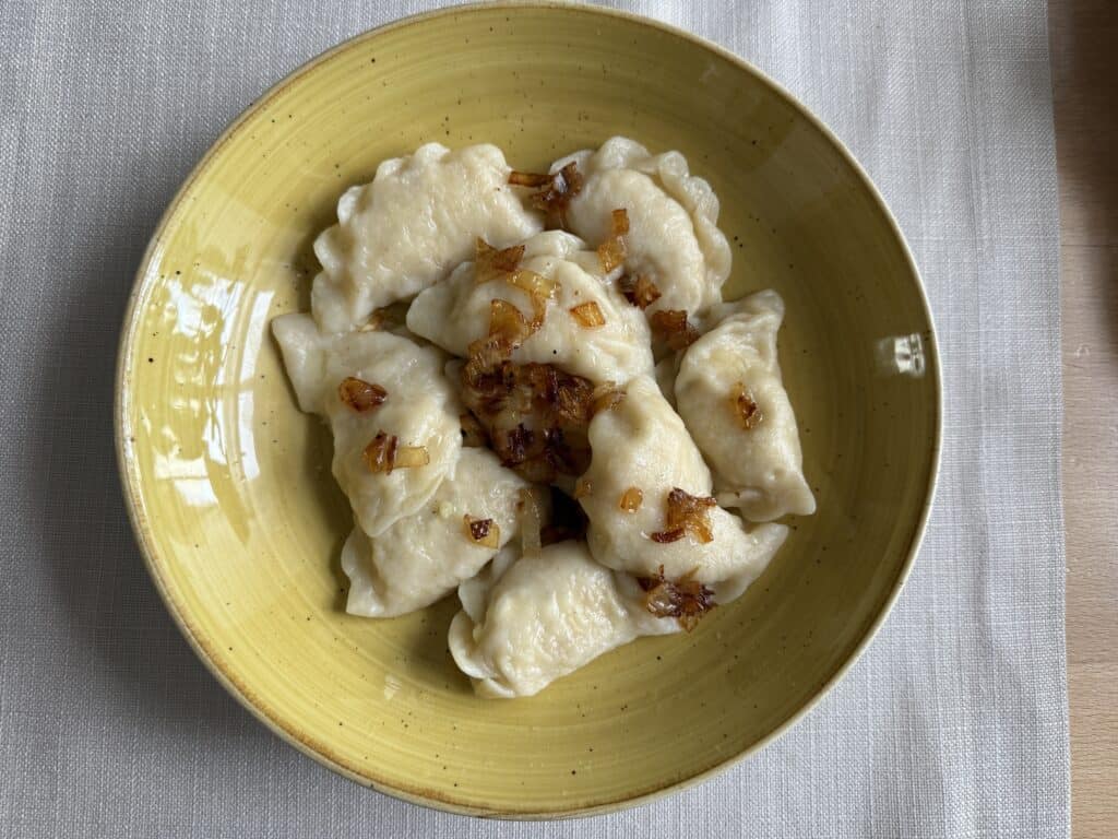 A yellow bowl with Pierogi Ruskie, topped with caramelized onions, rests on a white textured tablecloth.