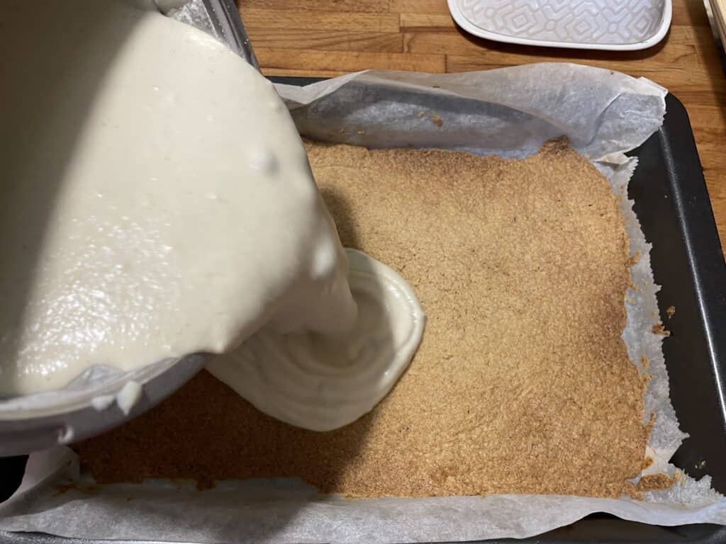 Pouring white vegan batter onto a pre-baked crust in a baking dish lined with parchment paper, likely for making a dessert.