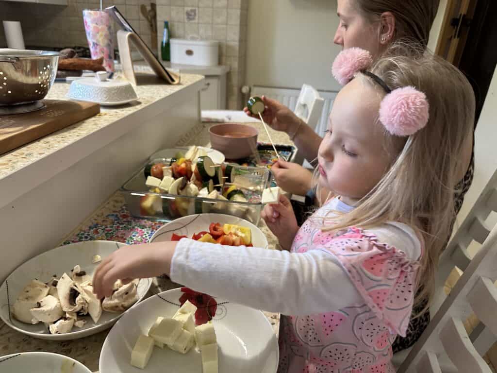 A young girl with pink earmuffs and an adult preparing Szaszłyki, a popular Polish cuisine dish, in a kitchen, with plates of vegetables and cheese on the counter.