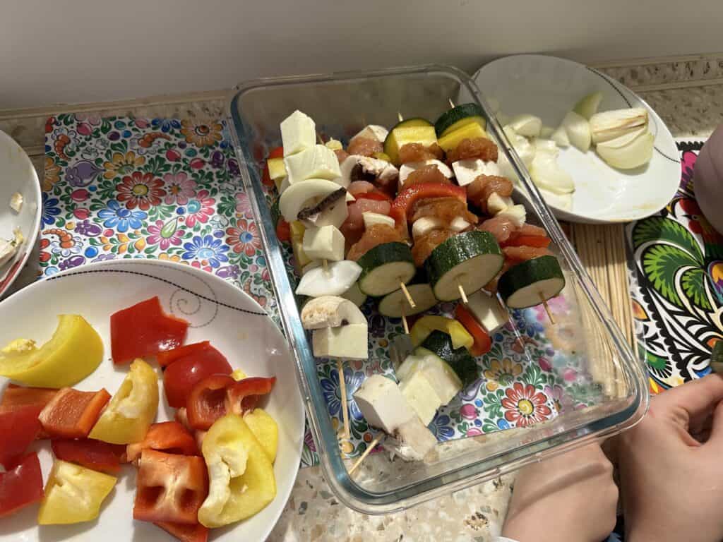 Ingredients for grilled skewers being prepared on a kitchen counter, featuring chopped peppers, zucchini, onions, and skewers with mixed vegetables and tofu.