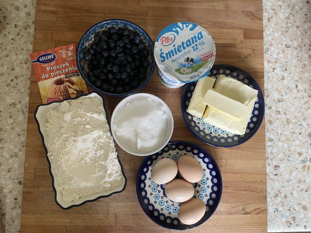 A Recipe display on the table: vibrant blueberries, baking powder, flour, and sugar mingle with rich sour cream, butter, and eggs atop patterned dishes. Perfect for crafting a traditional Polish Blueberry Babka (Babka Z Jagodami) that promises a delightful taste of heritage.