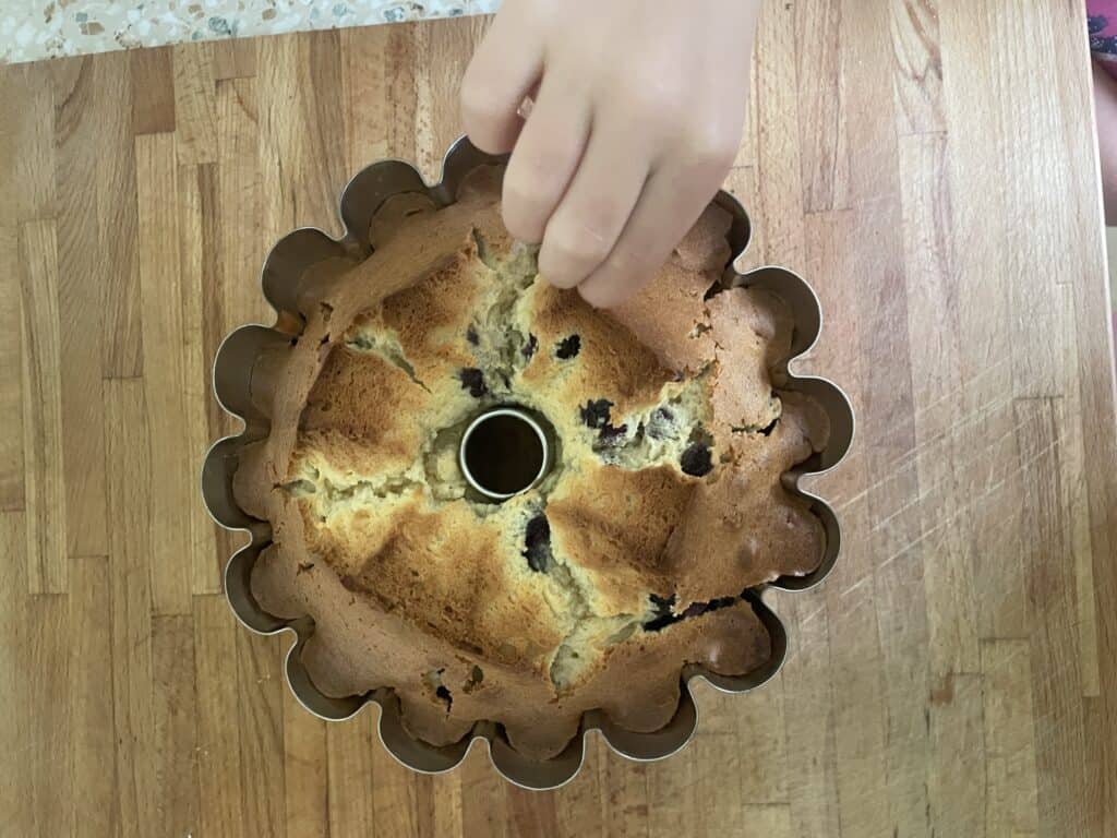 A hand sprinkles toppings on a freshly baked bundt cake with dark spots, reminiscent of a classic Polish Blueberry Babka Z Jagodami, resting invitingly on a wooden table.