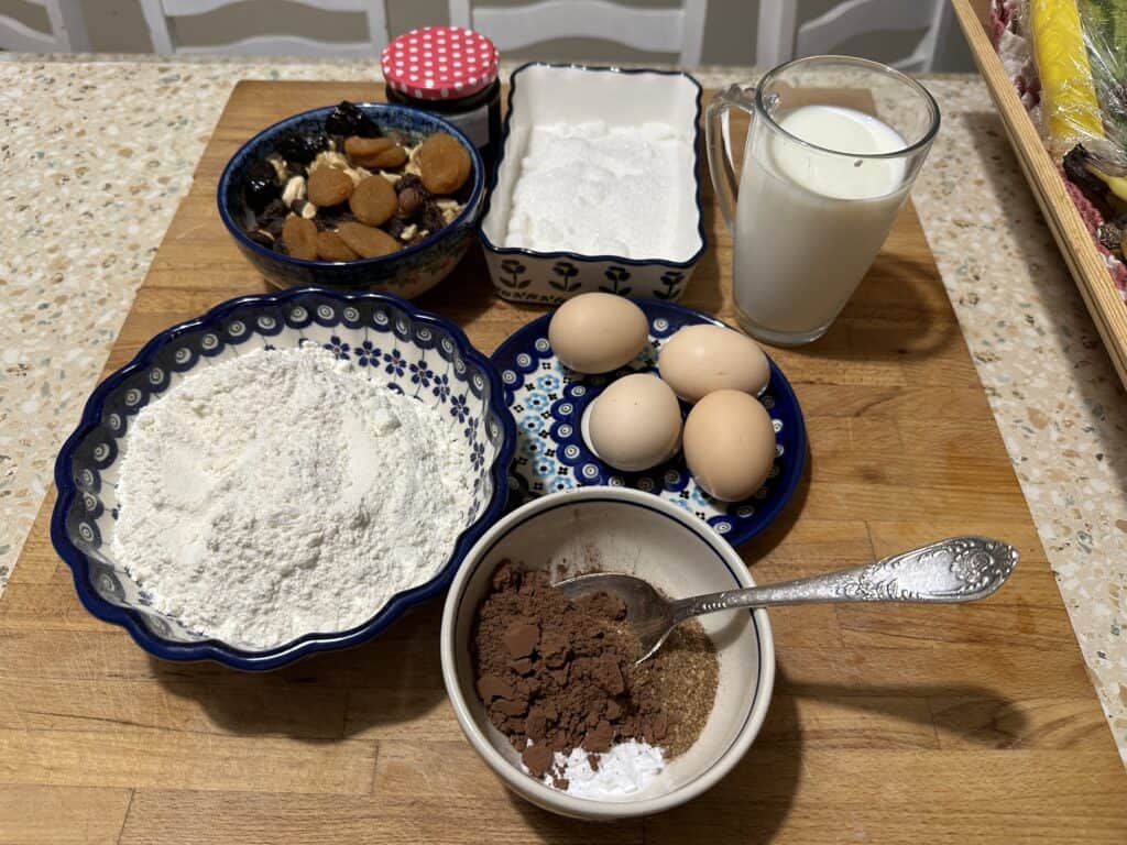 Baking ingredients on a wooden board for a delightful cake: flour, sugar, a bowl of nuts, eggs, cocoa powder, and a glass of milk.