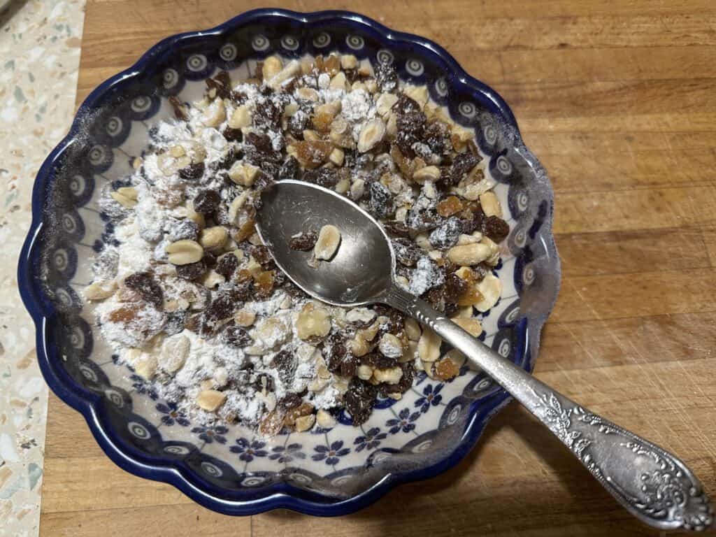 A patterned bowl filled with mixed nuts, raisins, and powdered sugar resembles a deconstructed gingerbread recipe, with an ornate silver spoon resting inside. The bowl sits on a wooden surface, inviting you to indulge in its sweet simplicity.
