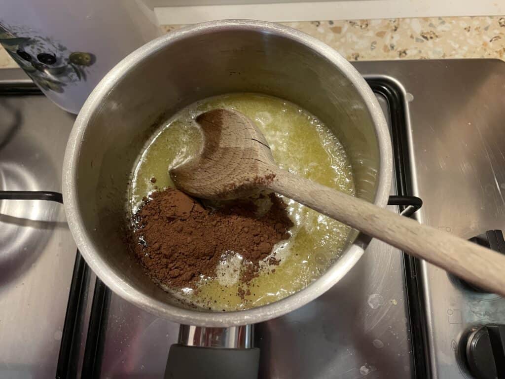 A wooden spoon stirs cocoa powder into melted butter on the stove, setting the stage for a quick gingerbread treat.