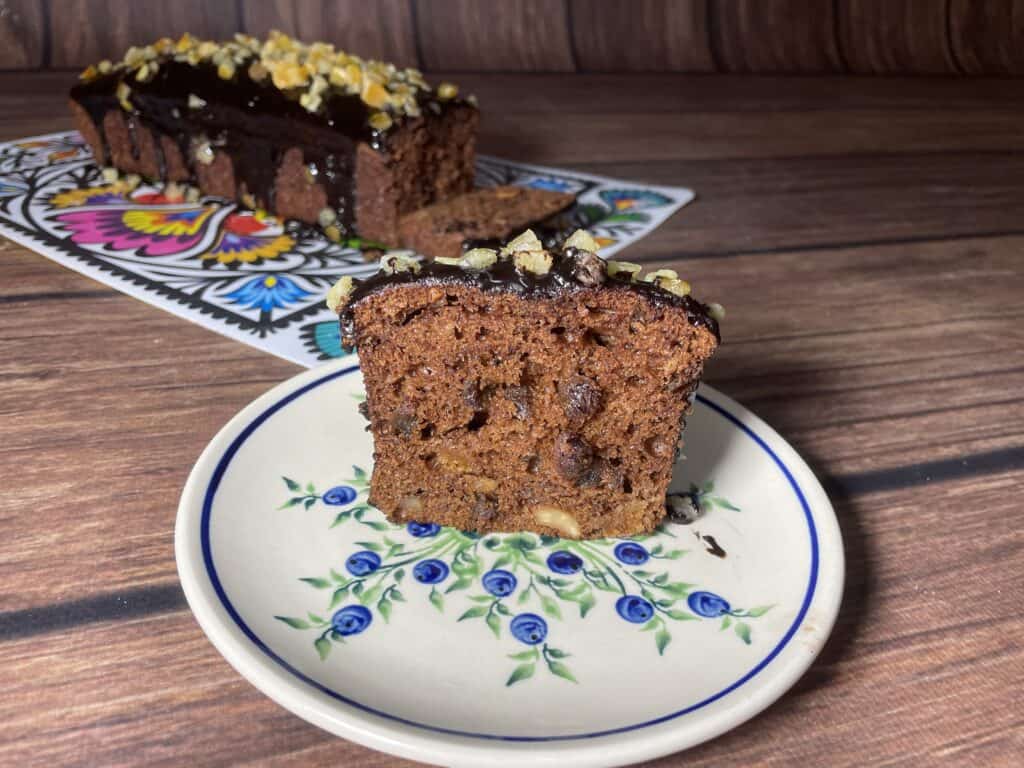 A slice of chocolate cake on a floral plate, with the rest in the background. Topped with chocolate icing and nuts, this delightful treat is a quick recipe for those who crave indulgence without the wait.