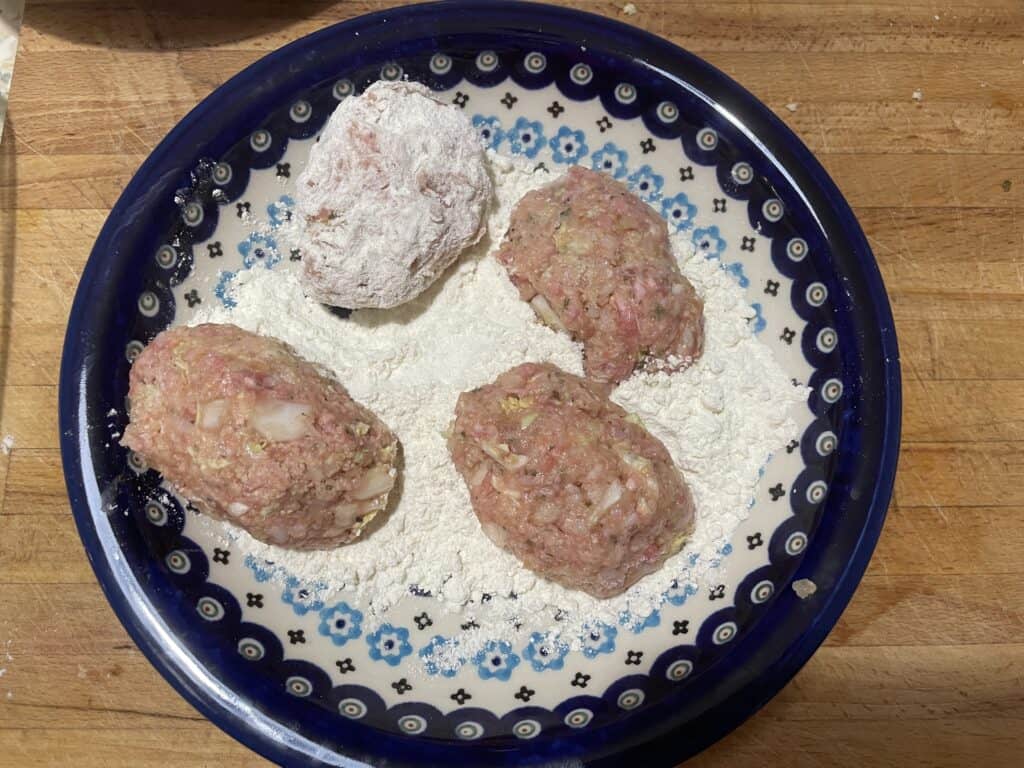 A blue-patterned bowl on a wooden surface holds four uncooked meatballs coated in flour, ready to become part of a quick recipe for Gołąbki Casserole.