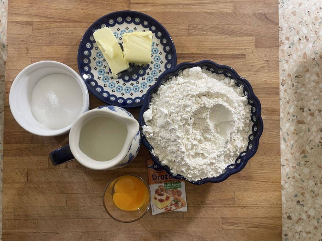Ingredients for baking: flour in a bowl, butter on a plate, sugar in a bowl, milk in a jug, egg yolks in a glass, and yeast packet on a wooden surface. These essentials set the stage for crafting traditional Rogale Świętomarcińskie, or Saint Martin's Croissants&mdash;a beloved Polish recipe.