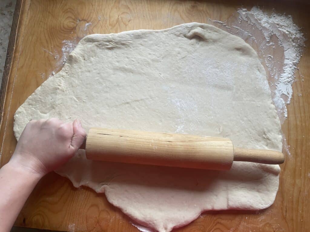 A hand uses a rolling pin to flatten dough for vegan pączki on a floured wooden surface.