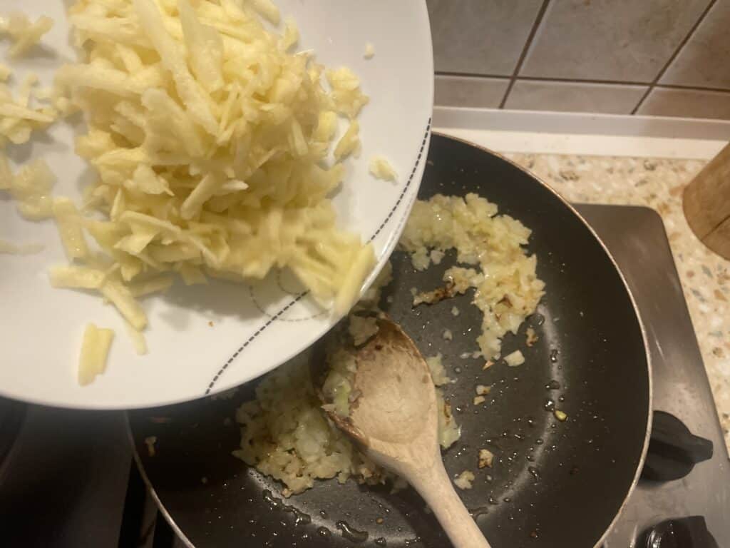 Grated apples are expertly incorporated with saut&eacute;ed onions in a frying pan using a wooden spoon, creating the base for a flavorful Polish-inspired Vegan Lard Spread, or Smalec Wegański.