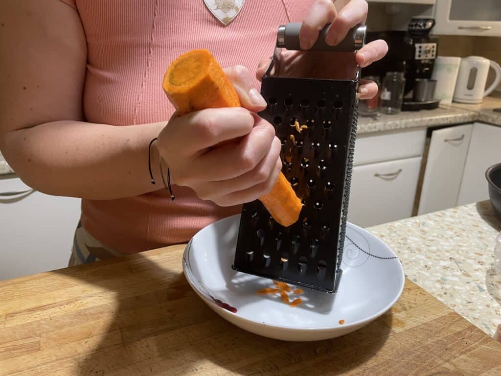 A person grating a carrot into a white bowl on the kitchen counter is preparing a hearty dish by adding vibrant color and fresh flavor to vegan kotlety mielone.