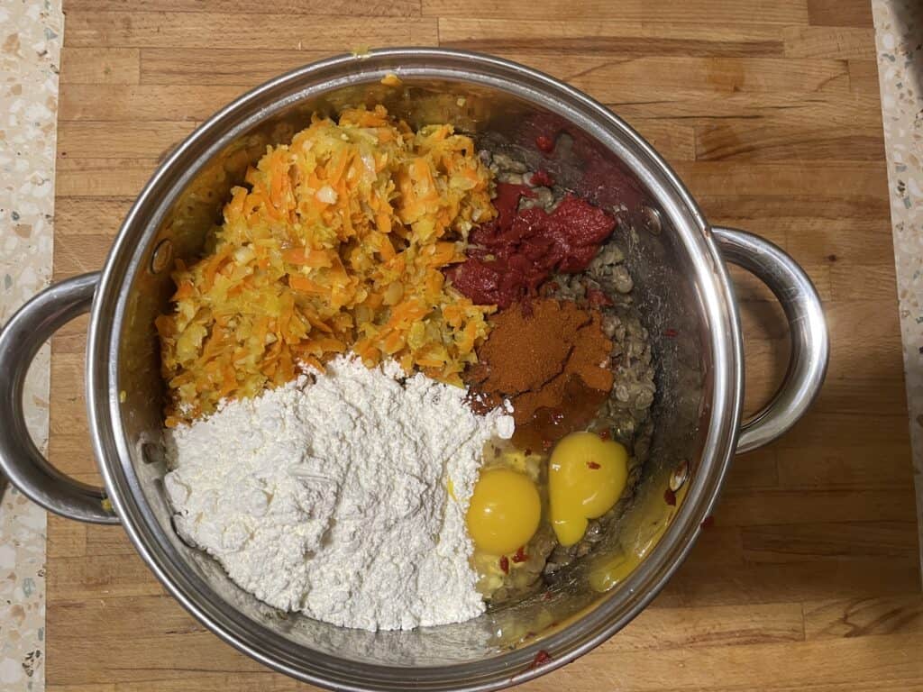 A mixing bowl with grated carrots, egg yolks, flour, spices, lentils, and tomato paste sits on a wooden surface next to other shredded vegetables. Perfect for crafting vegan kotlety mielone.