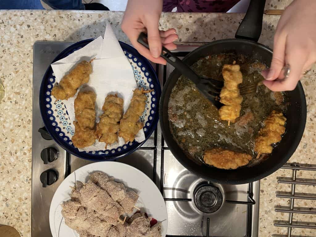 Person frying breadcrumb-coated skewers in a pan for a delicious city chicken recipe, with golden-brown pieces on a paper towel-lined plate and uncooked ones ready on another plate.