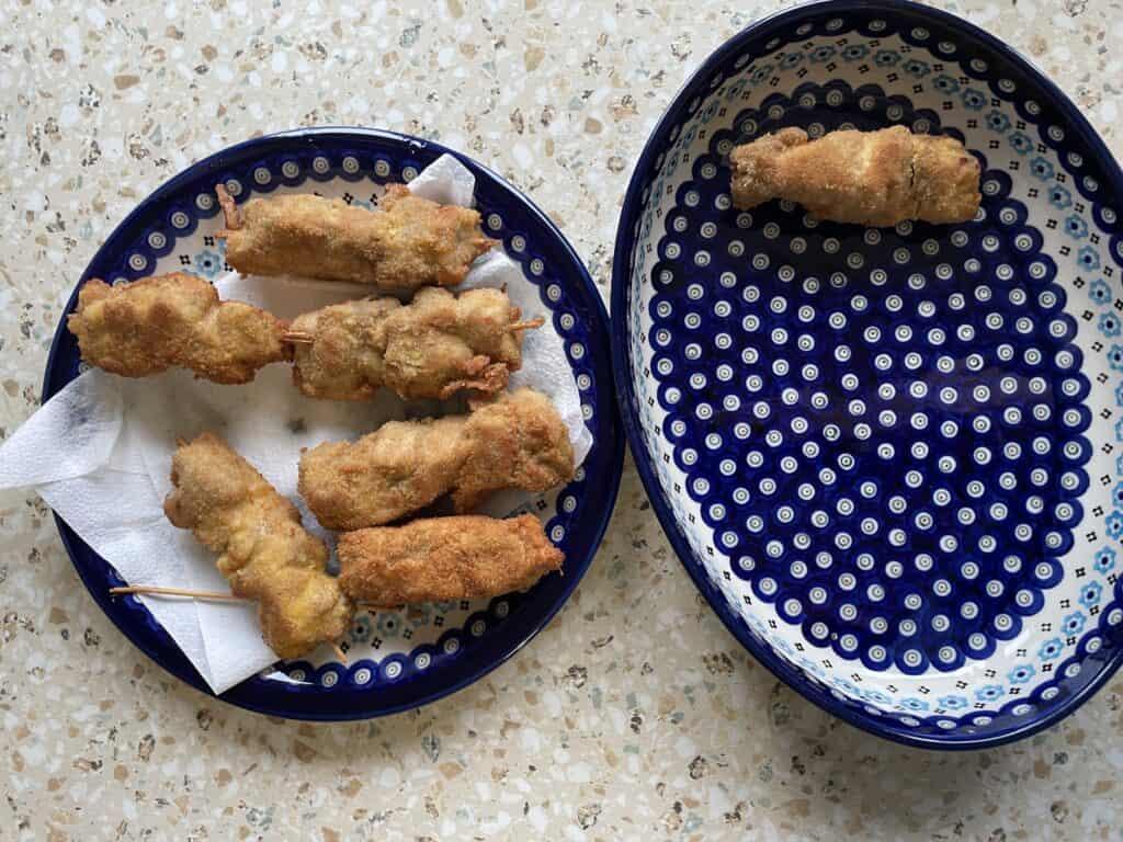 Two blue-patterned plates rest on a speckled surface; one holds several breaded skewers on a paper towel, the other features a single city chicken skewer. Perfect for trying out your grandma's City Chicken recipe at your next gathering.