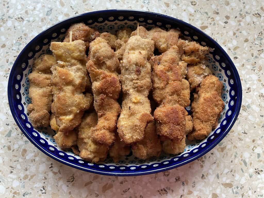 A plate of breaded and fried skewered meat, reminiscent of a city chicken recipe, is artfully arranged in a blue decorative bowl on a speckled countertop.