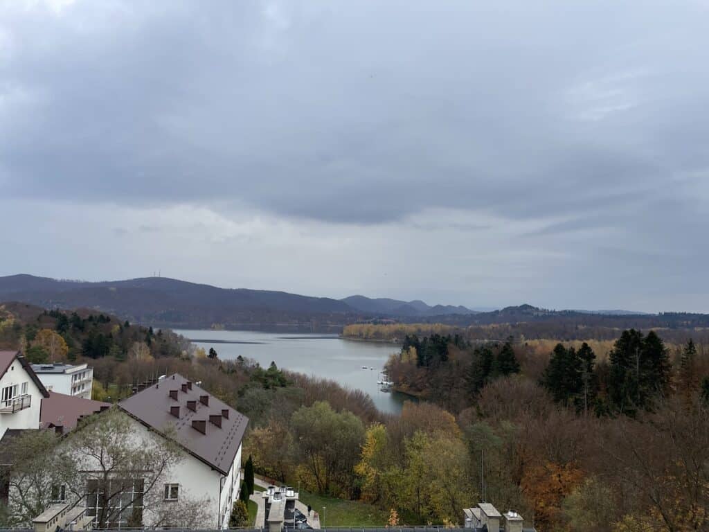 Experience the beauty of autumn in Poland with this view of a river or lake surrounded by trees with golden foliage, red-roofed buildings in the foreground, and distant hills beneath a cloudy sky.