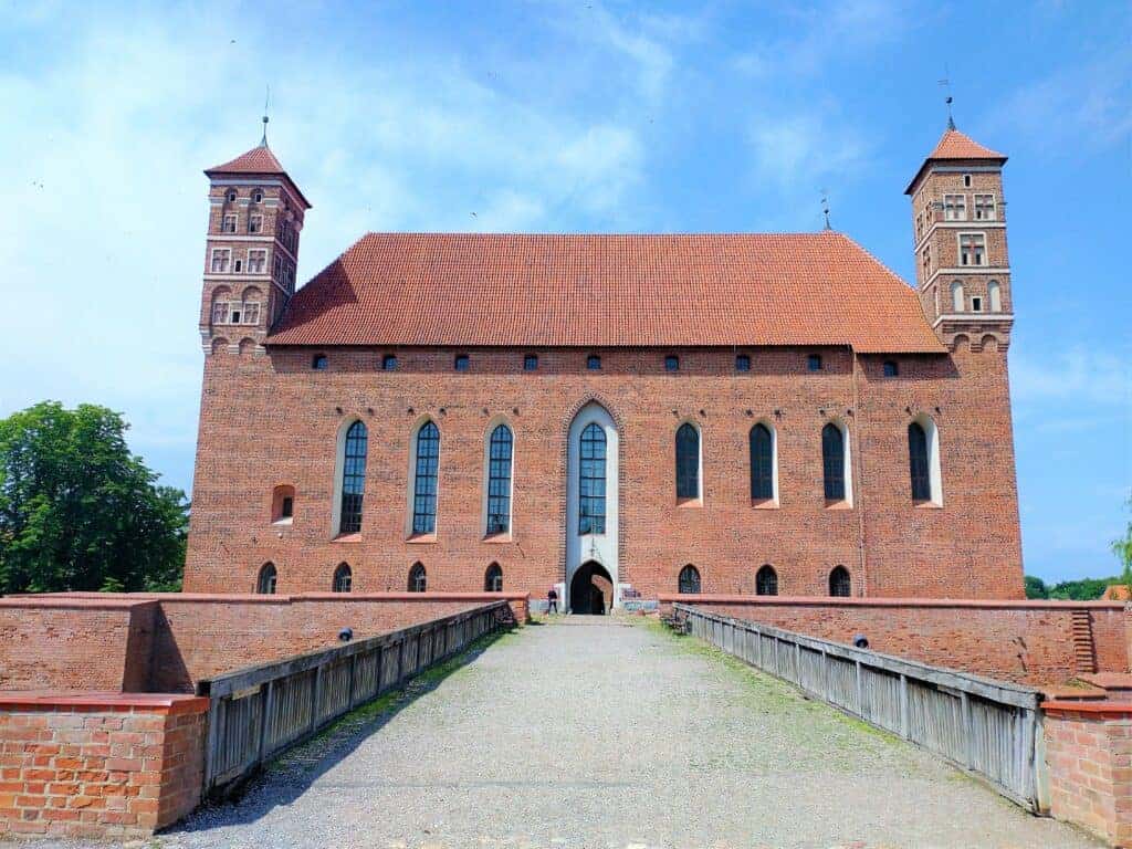 Large brick medieval castle with two square towers, arched windows, and a pitched red roof, viewed from a stone bridge under a blue sky&mdash;one of the must-see castles in Poland.