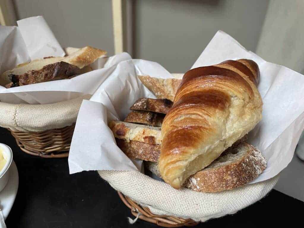 Two wicker baskets lined with white paper hold assorted breads, including a croissant and rustic loaf slices&mdash;an inviting display for the Best Breakfast Krak&oacute;w has to offer, all set on a dark surface.