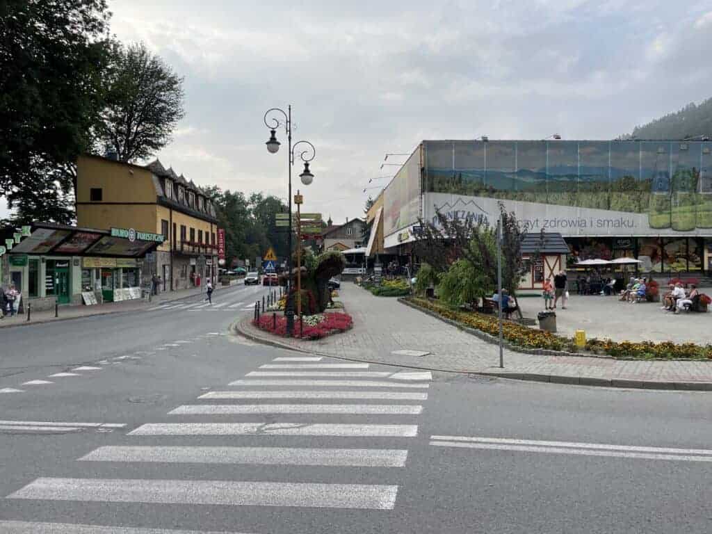 A crosswalk leads to a street corner with flowerbeds, shops, and people sitting outside a building on a cloudy day&mdash;an inviting scene that highlights Poland etiquette for enjoying public spaces respectfully.