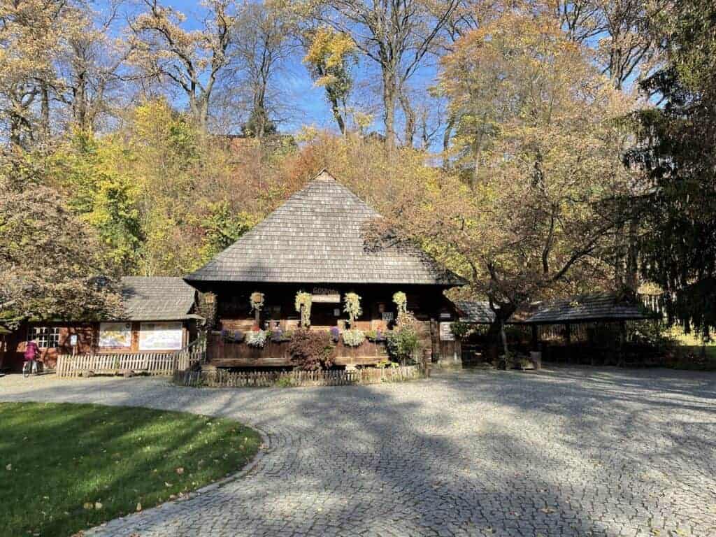 A rustic wooden house with a shingled roof sits among autumn trees, its flower boxes on the porch and cobblestone courtyard in front&mdash;capturing the cozy charm of autumn in Poland.