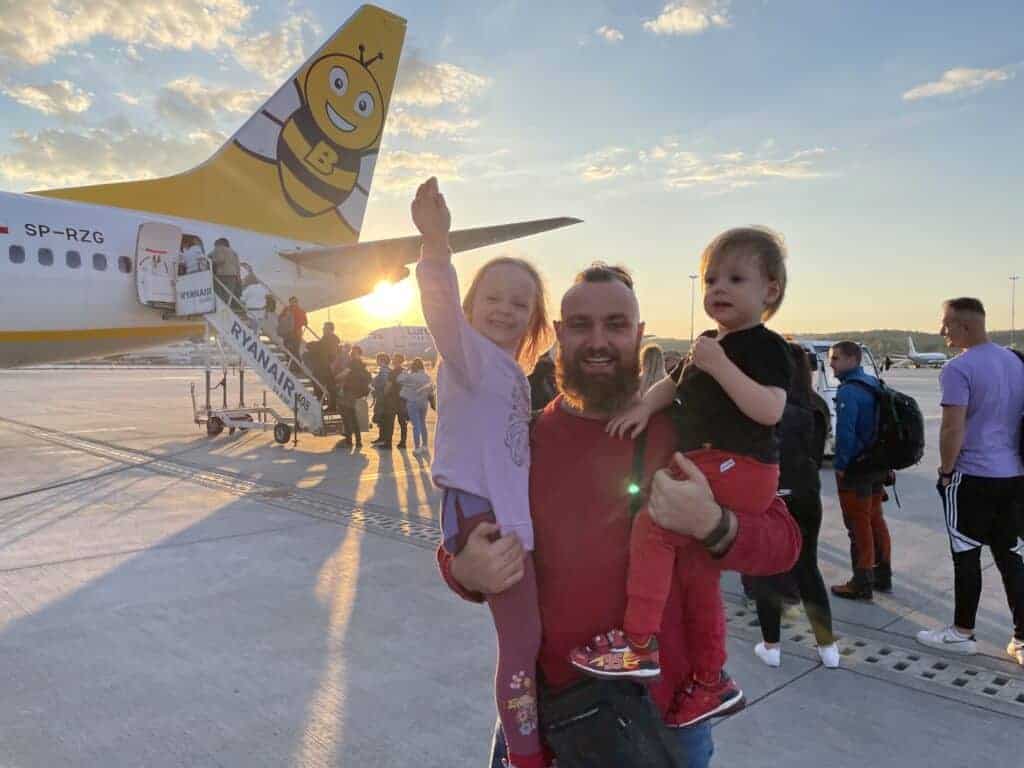 A man holds two children while standing on an airport tarmac near a Ryanair airplane; passengers boarding in the background are likely preparing for their flight to Poland.