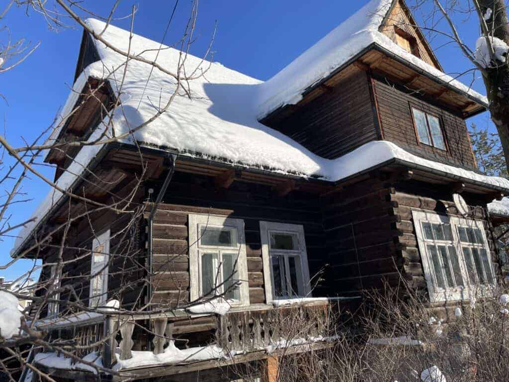 A wooden house with a snow-covered roof and windows, surrounded by bare branches under a clear blue sky&mdash;an idyllic scene capturing winter in Poland.