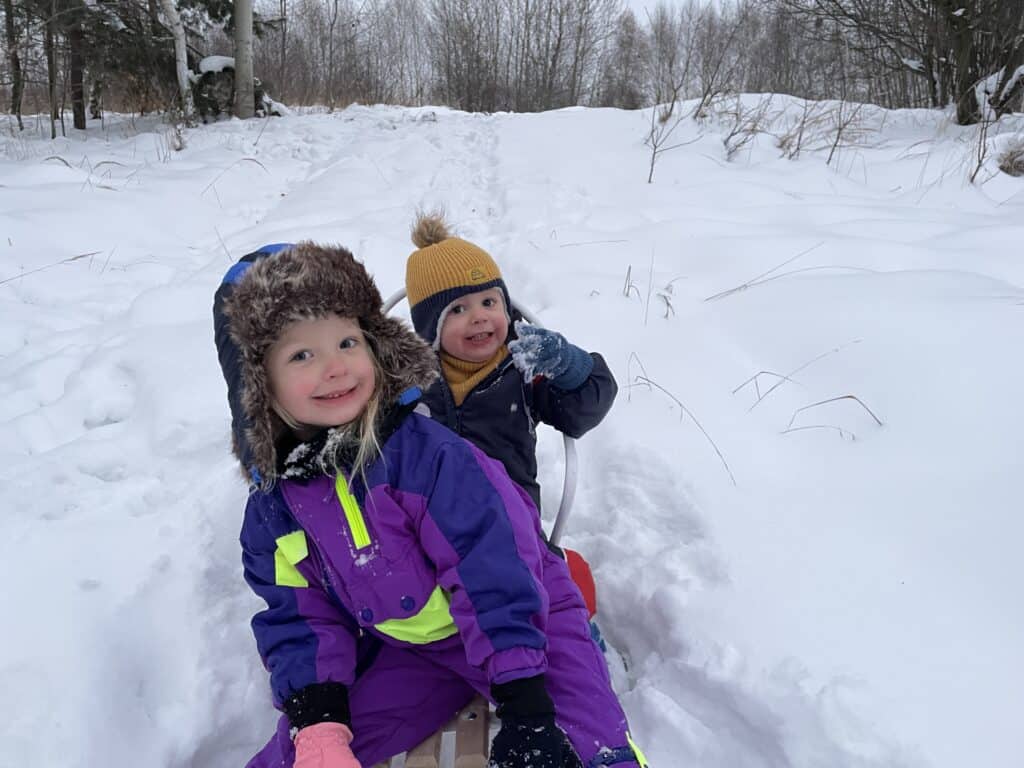 Two young children in winter clothing sit on a sled in a snowy outdoor setting, smiling at the camera with trees and snow-covered ground in the background&mdash;capturing the joy of winter in Poland.