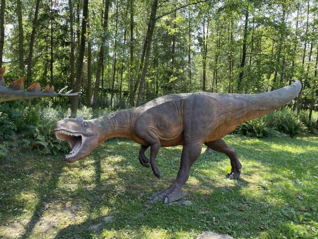 A life-sized dinosaur model with an open mouth stands outdoors on grass in Swietokrzyskie, Poland, surrounded by trees and greenery—one of the unique places to visit in the region.