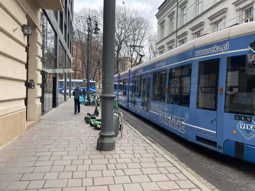A blue tram travels down a city street beside a sidewalk with parked electric scooters and a few people in the background, capturing the affordable charm of Poland travel cost for those exploring the city.