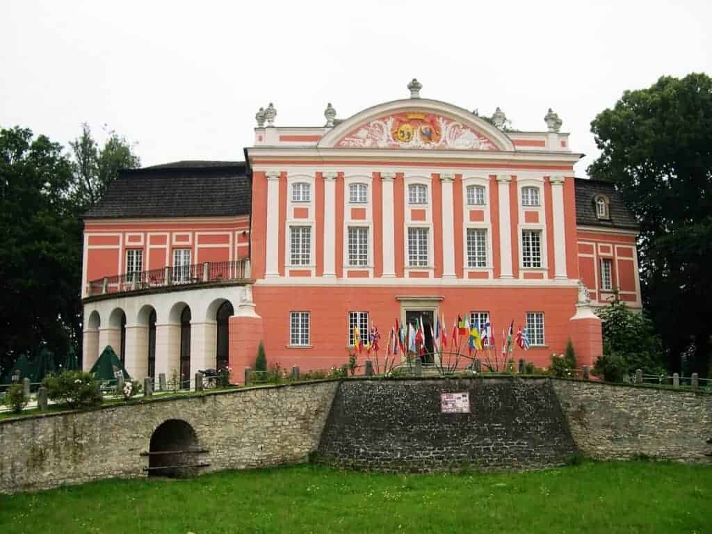 A large, ornate pink and white building with columns and multiple flags displayed at the entrance, set behind a stone wall and green lawn—one of the top places to visit in Swietokrzyskie, Poland.