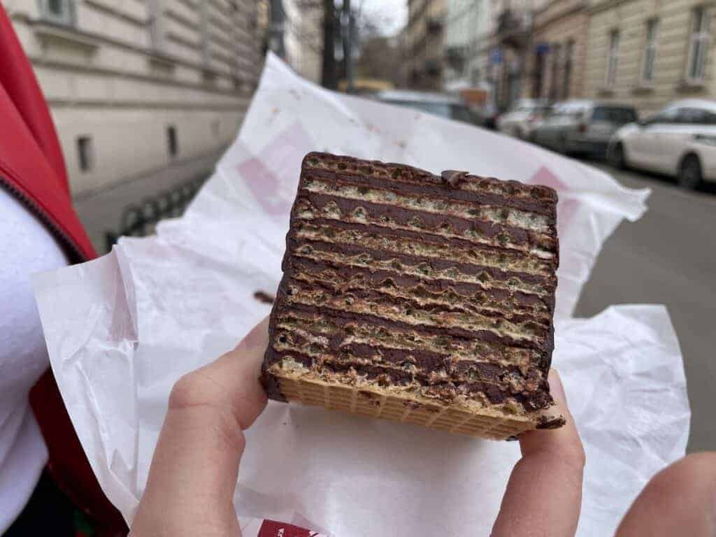 A person holding a large, multi-layered chocolate wafer dessert on a piece of white paper, perfect for anyone seeking the Best Breakfast Krak&oacute;w has to offer, with a street and parked cars in the background.