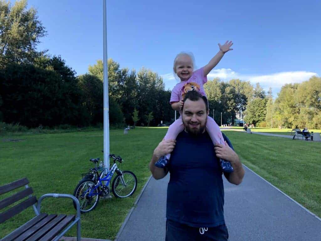 A man carries a smiling child on his shoulders in a park, with bicycles and benches nearby on a sunny day, capturing the joy of Polish Father's Day celebrated in Poland.