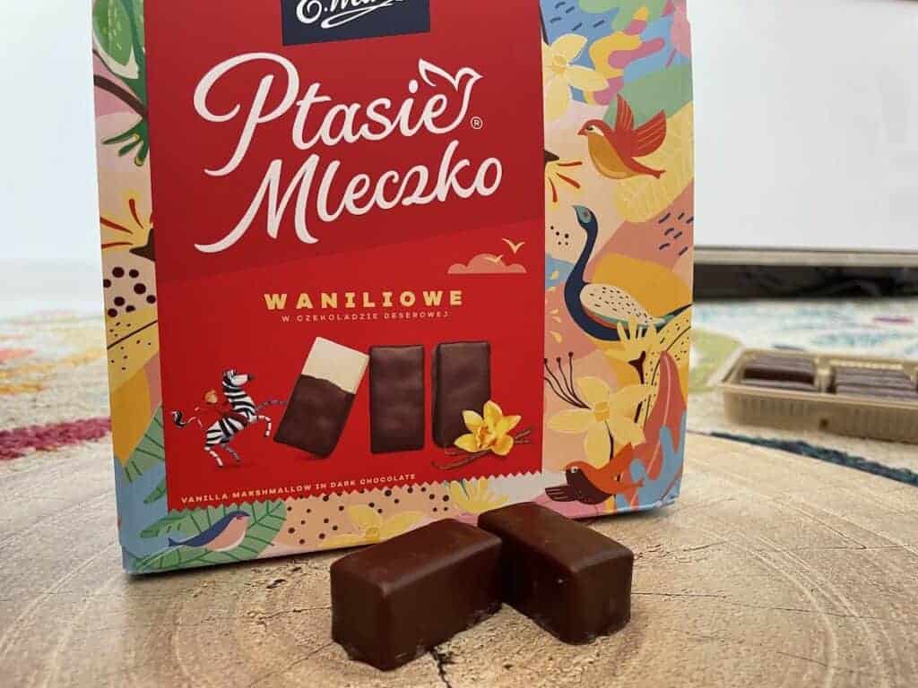 A colorful box of Ptasie Mleczko vanilla marshmallow candies in dark chocolate, a classic Polish souvenir, is shown with two candies in front and an open tray of sweets in the background.