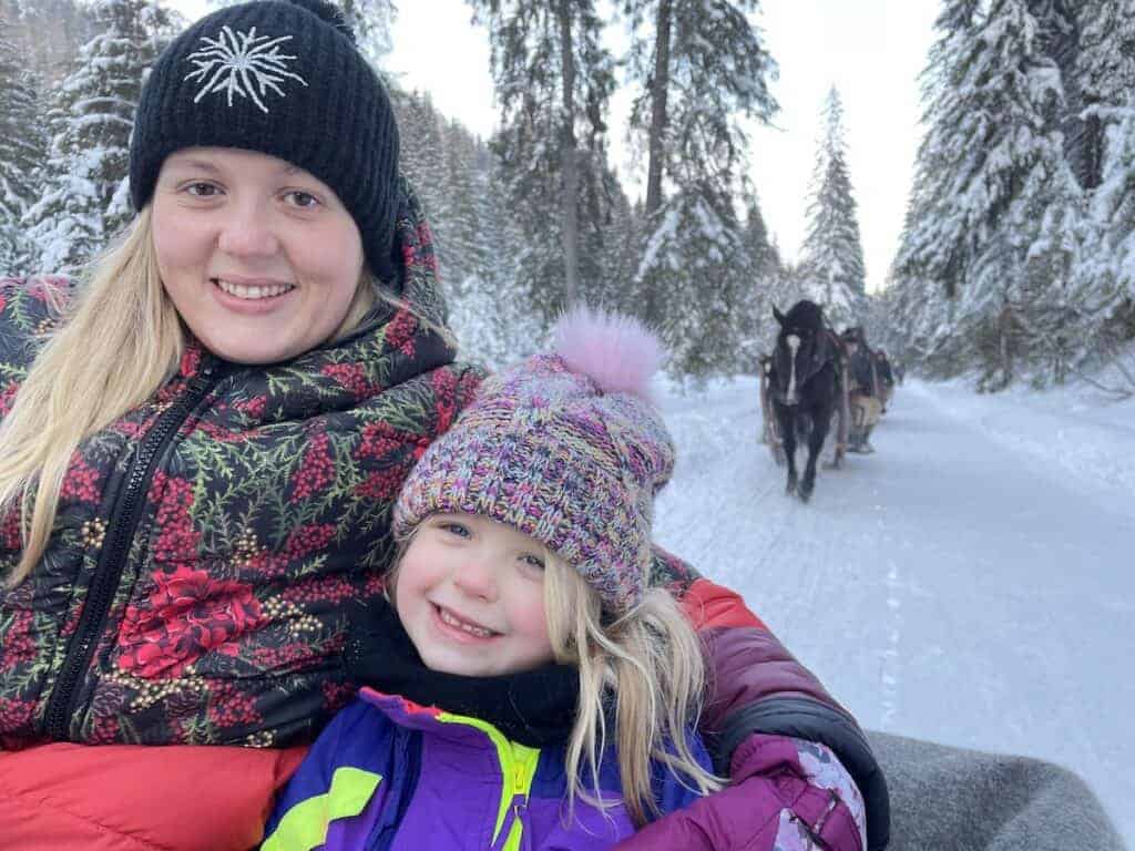 An adult and a child bundled in winter clothing ride in a horse-drawn sleigh through a snowy forest, enjoying the magic of snow in Poland, with another sleigh visible behind them.