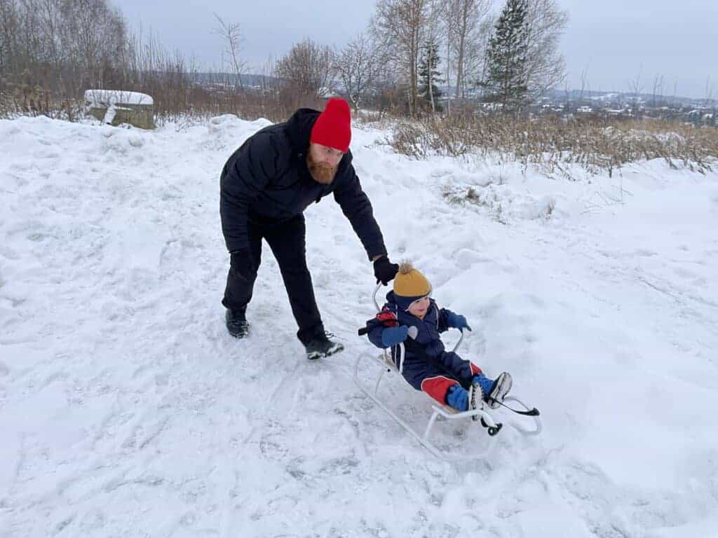 An adult pushes a child on a sled across a snowy landscape, with trees and an overcast sky in the background, capturing the joy of Zakopane winter.