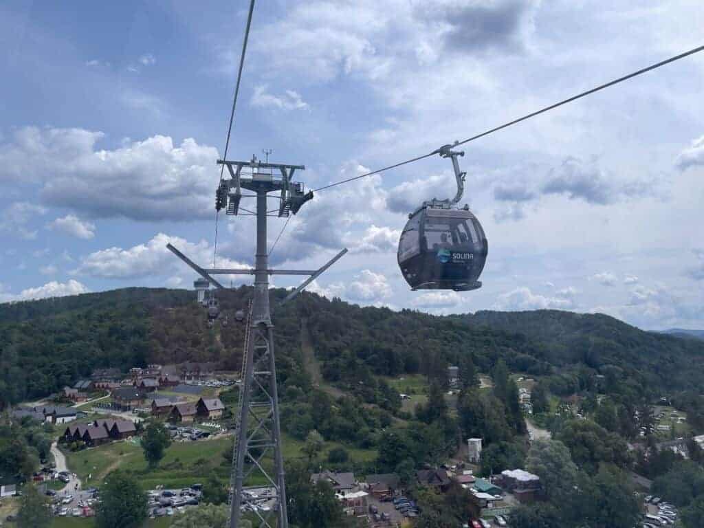 A gondola lift travels over a green, hilly landscape in Poland&rsquo;s Bieszczady Mountains, with a village and parked cars below under a partly cloudy sky&mdash;an insider&rsquo;s guide to scenic mountain views.