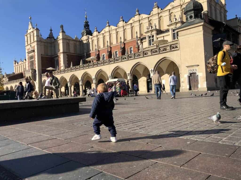 A small child stands on a cobblestone square in Krak&oacute;w in December, surrounded by people walking and pigeons near a historic building&mdash;capturing one of the many things to do during winter in the city.