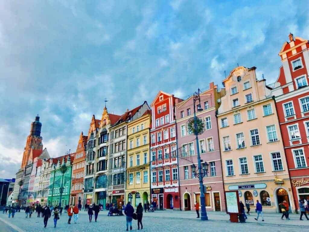 People walk along a cobblestone street lined with colorful, historic buildings under a partly cloudy sky, capturing the charm of Poland&rsquo;s historical landmarks.