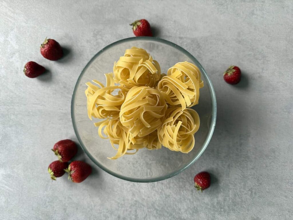 A glass bowl with six nests of uncooked fettuccine pasta is surrounded by fresh strawberries, evoking the classic Polish pasta dish Makaron z truskawkami—perfect inspiration for your next strawberries recipe.