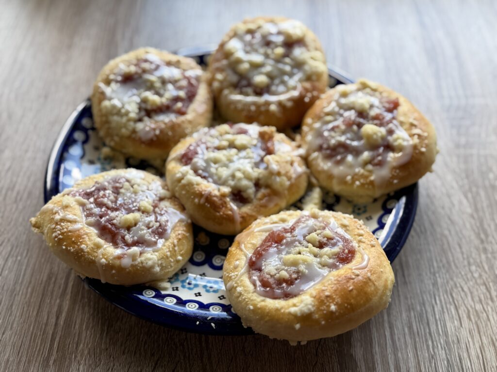 A plate with six round pastries, classic Drożdż&oacute;wki or Polish Apple Yeast Buns, topped with fruit jam and crumbled streusel, resting on a wooden surface&mdash;an easy recipe for a delightful treat.