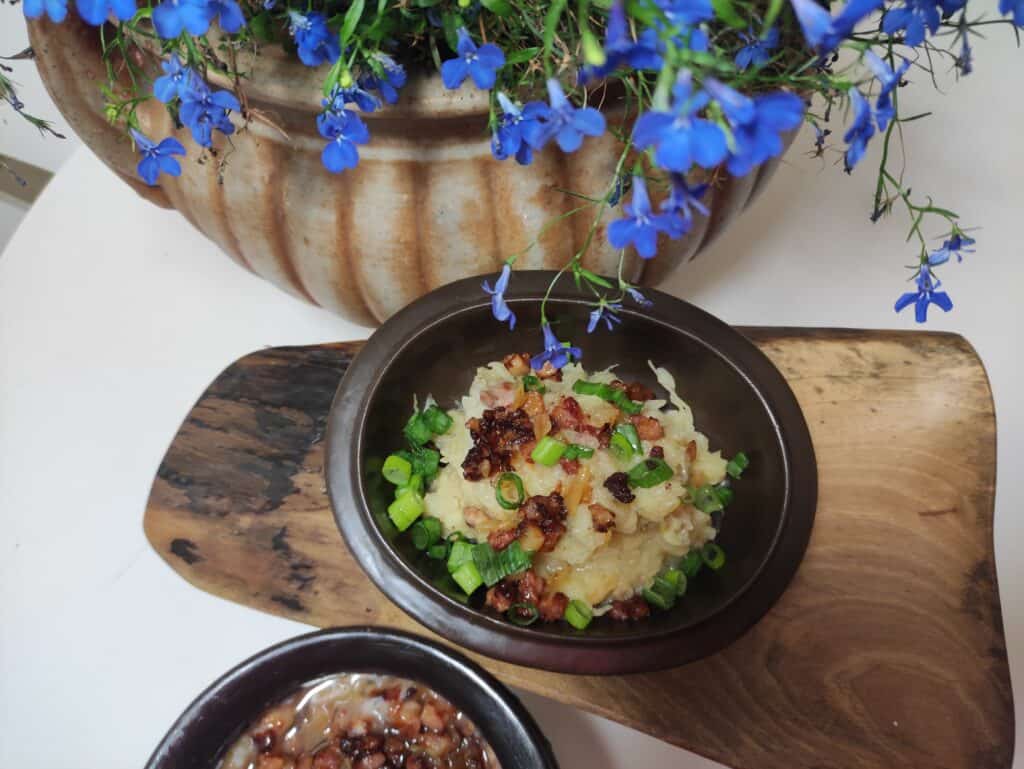 A bowl of rice topped with green onions and crispy bits, placed on a wooden board. A pot with blue flowers is in the background, evoking the simplicity of a traditional Polish sauerkraut recipe.