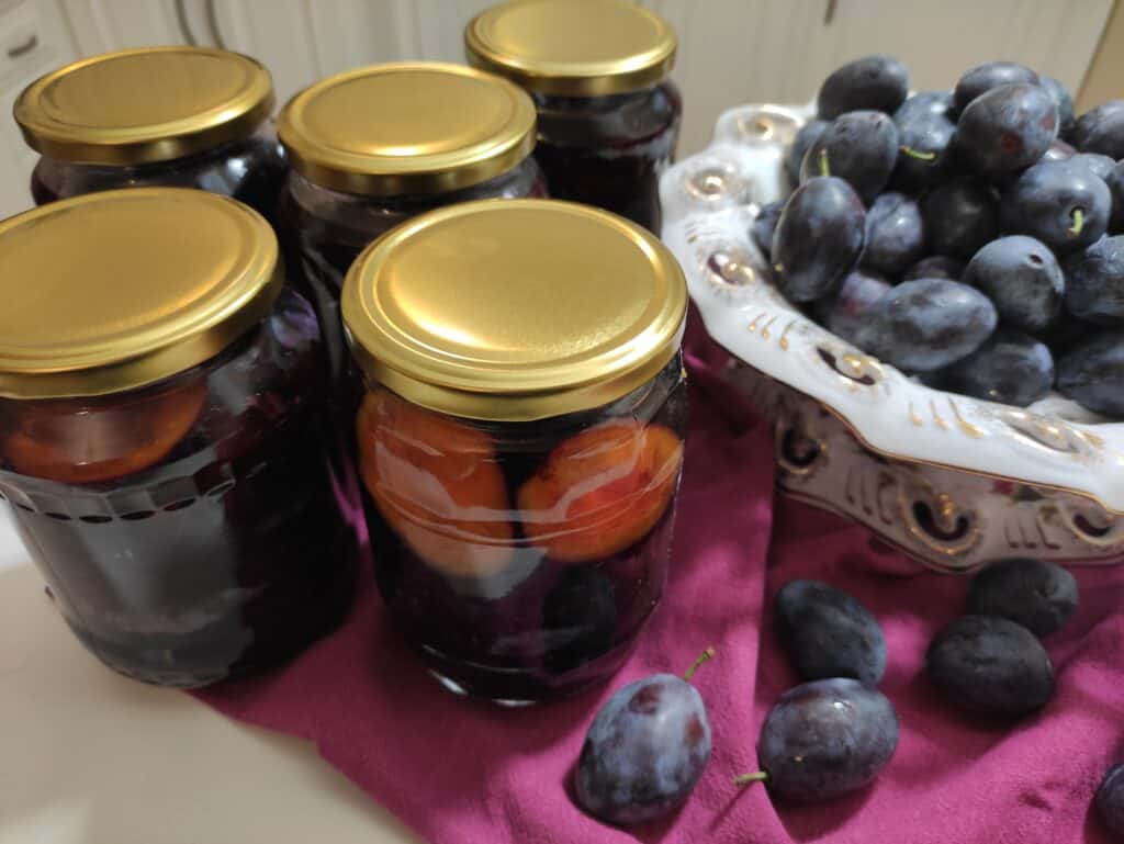 Five jars of preserved plums with gold lids are on a purple cloth next to a bowl of fresh plums, showcasing the splendid Śliwki W Occie, a Polish Vinegar Pickled Plums recipe.